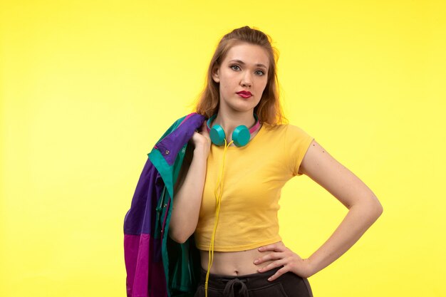 Une vue de face jeune femme moderne en chemise jaune pantalon noir et veste colorée avec des écouteurs colorés posant