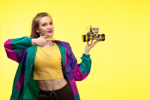 Une vue de face jeune femme moderne en chemise jaune pantalon noir et veste colorée avec holding toy car posant