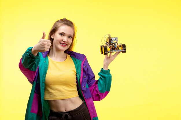 Une vue de face jeune femme moderne en chemise jaune pantalon noir et veste colorée tenant une petite voiture posant une expression heureuse