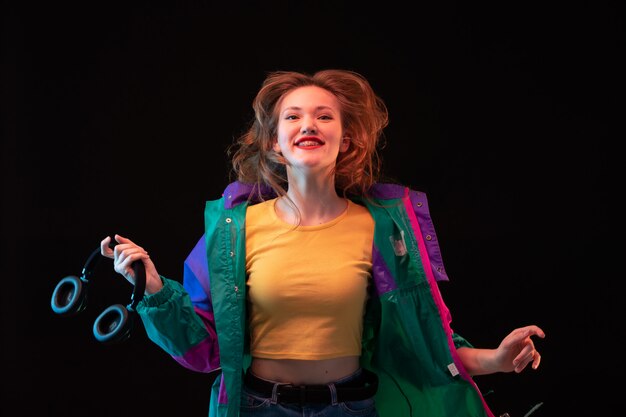 Une vue de face jeune femme moderne en manteau coloré orange t-shirt avec des écouteurs noirs posant heureux sur le fond noir de la danse de la mode moderne