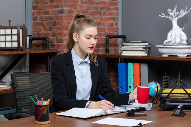 Vue de face d'une jeune femme occupée assise à une table et tenant une tasse rouge écrivant sur un document au bureau