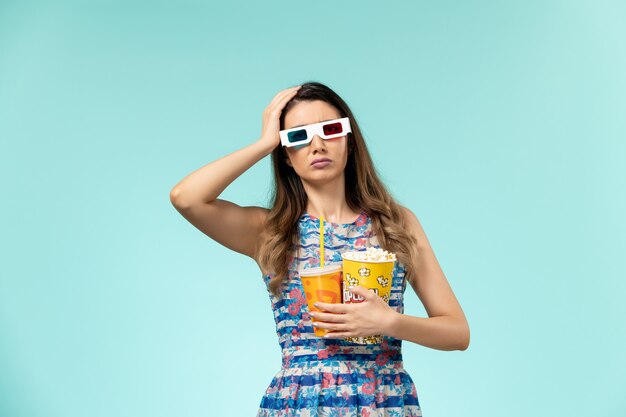 Vue de face jeune femme avec paquet de pop-corn et boire dans des lunettes de soleil sur une surface bleue