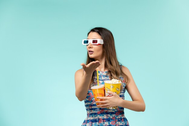 Vue de face jeune femme avec paquet de pop-corn et boire dans des lunettes de soleil sur une surface bleue