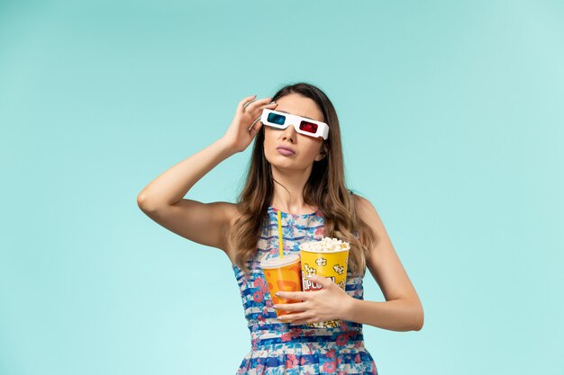 Vue de face jeune femme avec paquet de pop-corn et boire dans des lunettes de soleil sur une surface bleue