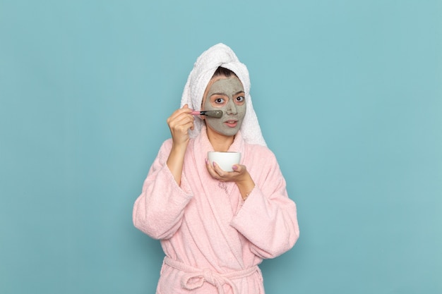 Vue de face jeune femme en peignoir rose après la douche, appliquer la crème sur le mur bleu de l'eau de beauté douche propre