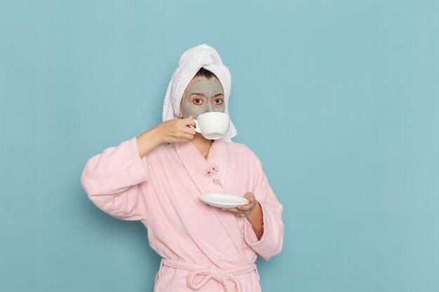 Vue de face jeune femme en peignoir rose après la douche, boire du café sur le mur bleu clair beauté eau propre douche selfcare