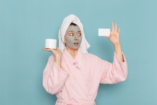 Vue de face jeune femme en peignoir rose après la douche avec masque sur son visage tenant la carte sur le mur bleu douche nettoyage crème auto-soin de beauté