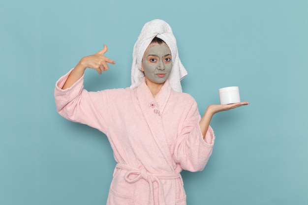 Vue de face jeune femme en peignoir rose après la douche avec masque sur son visage tenant la crème sur le mur bleu douche nettoyage beauté crème auto-soin