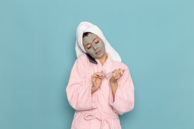 Vue de face jeune femme en peignoir rose après la douche parler au téléphone sur le bureau bleu clair de l'eau de beauté douche propre