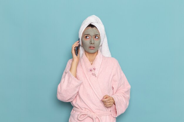 Vue de face jeune femme en peignoir rose après la douche parler au téléphone sur le mur bleu clair de l'eau de beauté douche propre