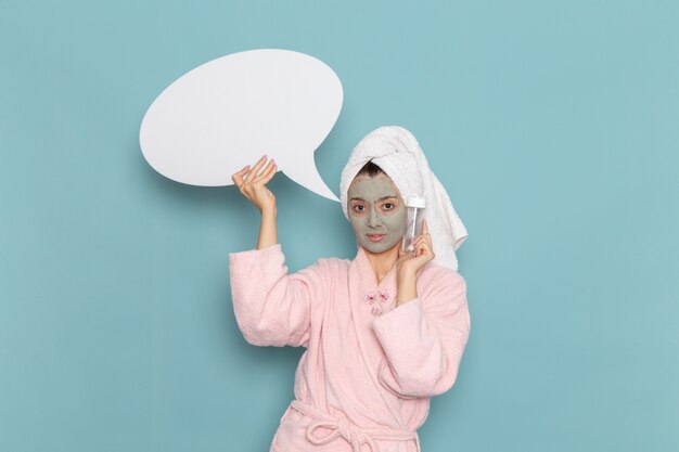 Vue de face jeune femme en peignoir rose après la douche tenant signe et vaporiser sur le mur bleu beauté eau crème auto-soin douche salle de bains