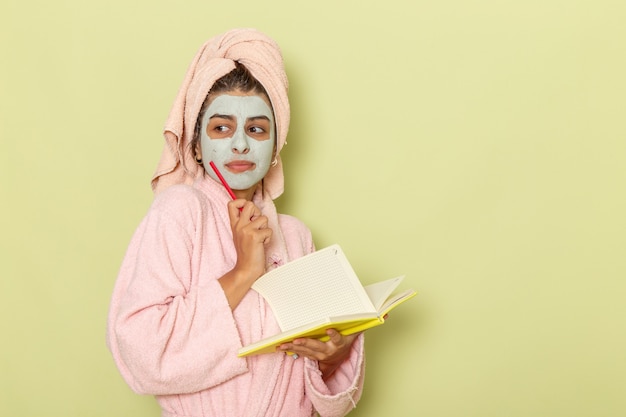 Vue de face jeune femme en peignoir rose en écrivant des notes sur une surface vert clair