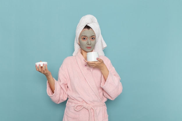 Vue de face jeune femme en peignoir rose avec masque sur son visage tenant la crème sur le bureau bleu douche nettoyage crème auto-soin de beauté