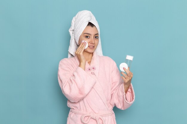 Vue de face jeune femme en peignoir rose nettoyer son visage de maquillage sur bureau bleu nettoyage douche crème auto-soin