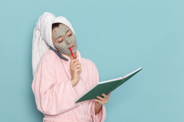 Vue de face jeune femme en peignoir rose parler au téléphone et lire le cahier sur bureau bleu nettoyage douche crème auto-soin