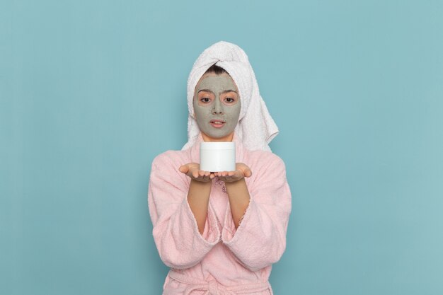 Vue de face jeune femme en peignoir rose tenant la crème sur le mur bleu clair douche nettoyage beauté crème auto-soins