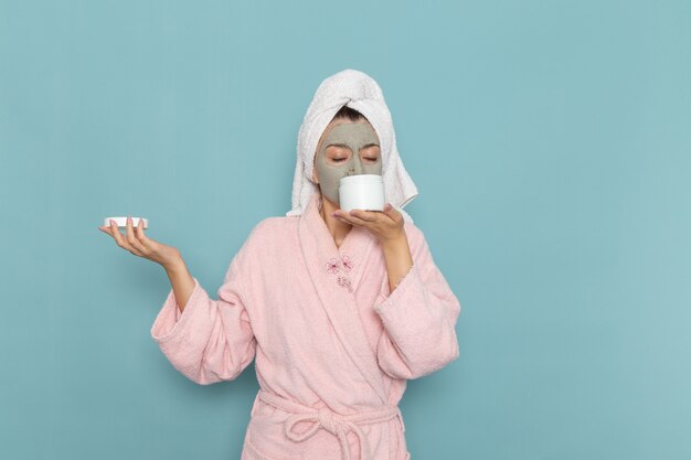 Vue de face jeune femme en peignoir rose tenant la crème sur le mur bleu crème de bain d'eau de beauté douche auto-soins