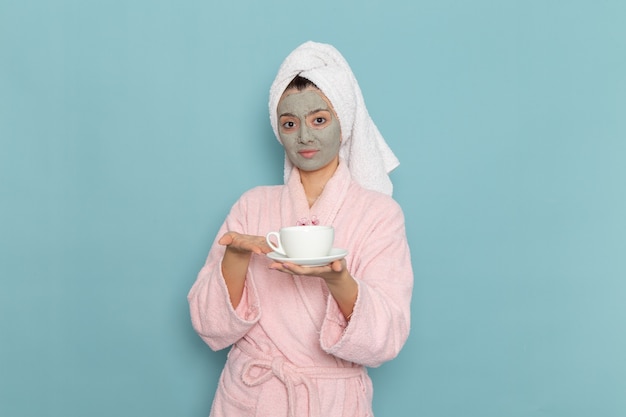 Vue de face jeune femme en peignoir rose tenant une tasse de café sur le mur bleu nettoyage douche crème auto-soin