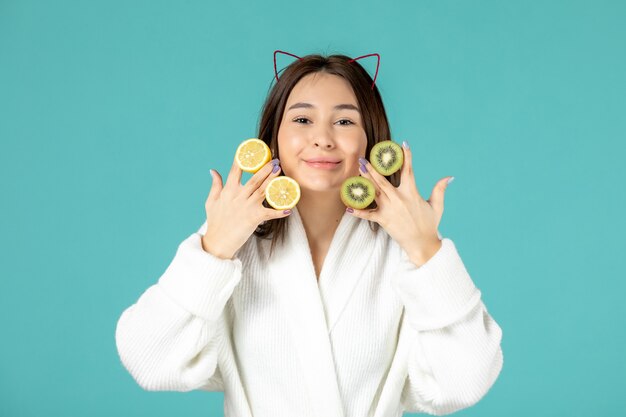 vue de face jeune femme en peignoir tenant des tranches de citrons et de kiwis sur fond bleu