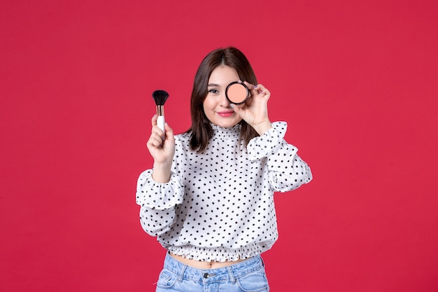 Vue de face d'une jeune femme avec pompon et poudre faisant du maquillage sur un mur rouge