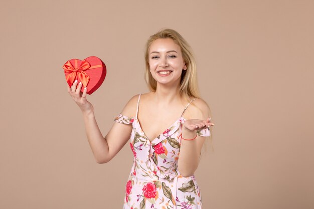 Vue de face d'une jeune femme posant avec un présent en forme de coeur rouge sur un mur marron