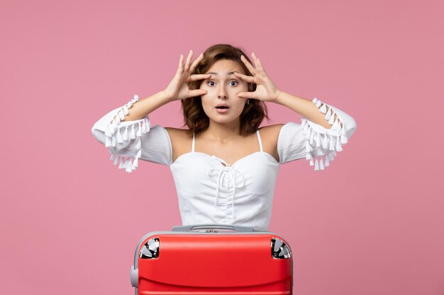 Vue de face d'une jeune femme posant avec un sac de vacances rouge sur un mur rose clair