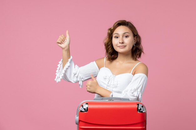 Vue de face d'une jeune femme posant avec un sac de vacances rouge sur le mur rose