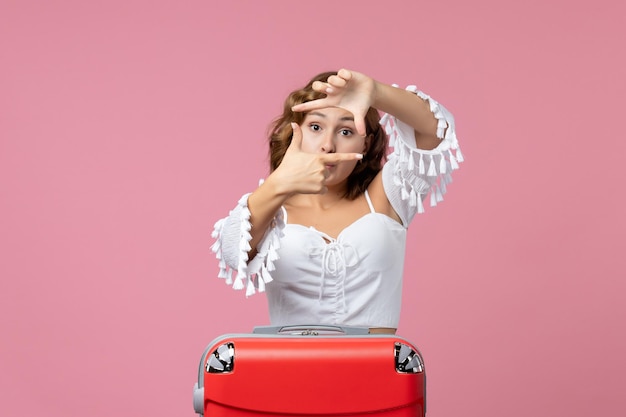 Vue de face d'une jeune femme posant avec un sac de vacances rouge sur le mur rose