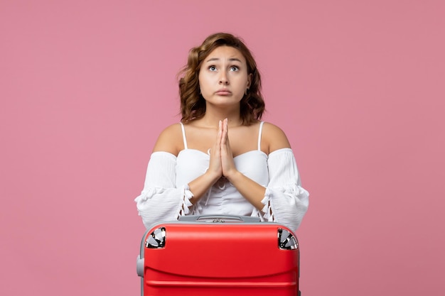 Vue de face d'une jeune femme priant avec un sac de vacances rouge sur un mur rose