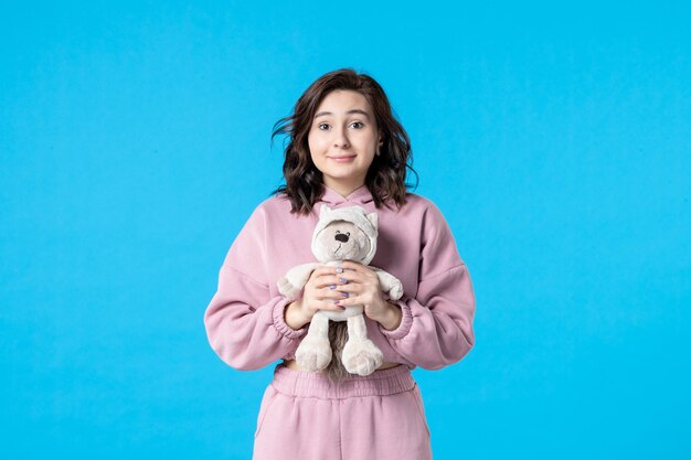 Vue de face jeune femme en pyjama rose avec petit ours en peluche sur bleu