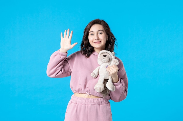 Vue de face jeune femme en pyjama rose avec petit ours en peluche sur bleu