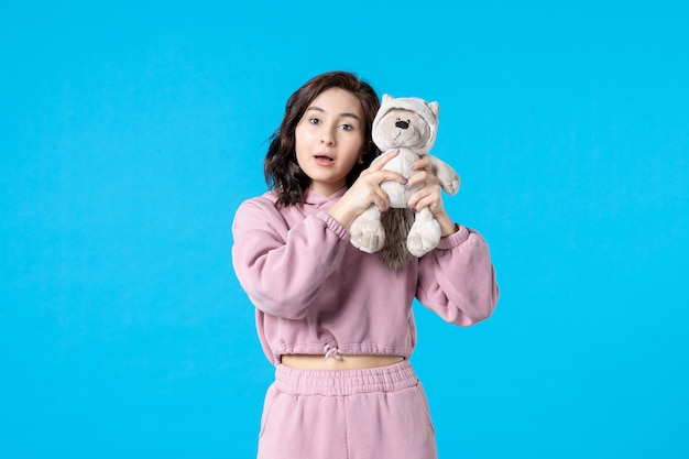 Vue de face jeune femme en pyjama rose avec petit ours en peluche sur bleu