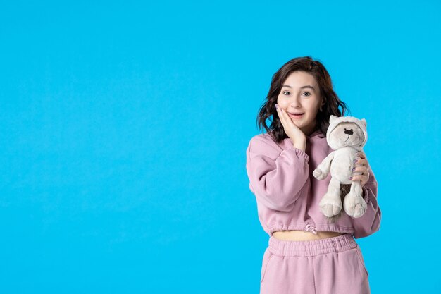 Vue de face jeune femme en pyjama rose avec petit ours en peluche sur bleu