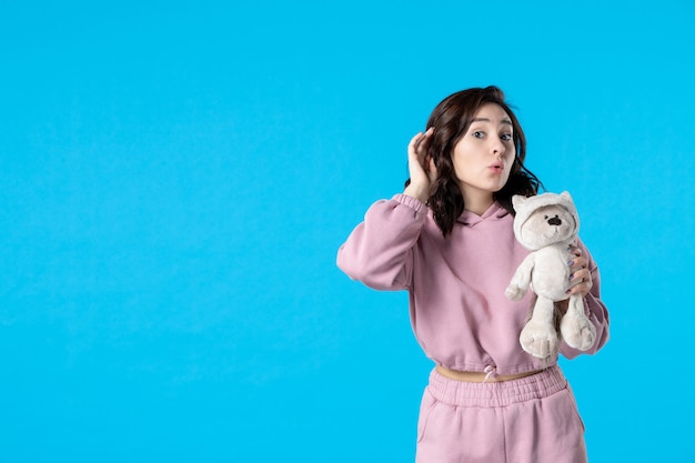 Vue de face jeune femme en pyjama rose avec petit ours en peluche sur bleu