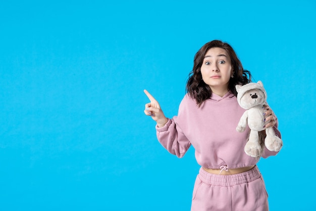 Vue de face jeune femme en pyjama rose avec petit ours en peluche sur bleu