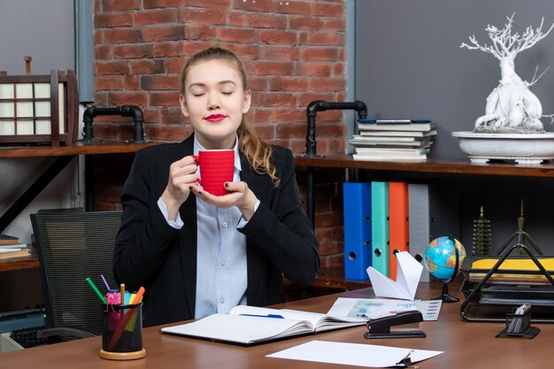 Vue de face d'une jeune femme rêveuse assise à une table et tenant une tasse rouge au bureau