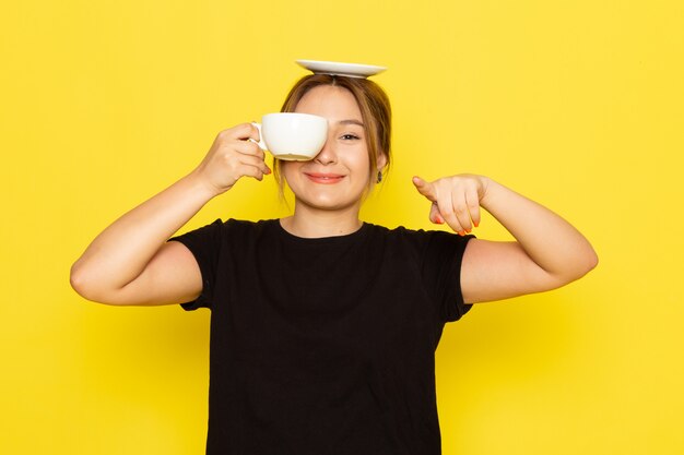 Une vue de face jeune femme en robe noire, boire du café et souriant sur jaune