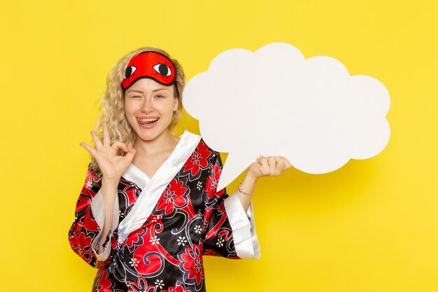 Vue de face jeune femme en robe de nuit masque pour les yeux tenant un énorme panneau blanc sur le mur jaune sommeil nuit couleur modèle