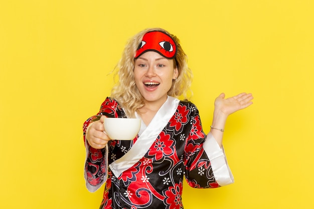 Vue de face jeune femme en robe de nuit et portant un masque pour les yeux se préparant à dormir tenant une tasse de café sur le mur jaune sommeil modèle féminin lit de nuit