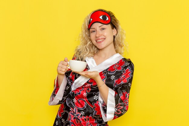 Vue de face jeune femme en robe de nuit et portant un masque pour les yeux se préparant à dormir tenant une tasse de café souriant sur un bureau jaune sommeil modèle féminin lit de nuit