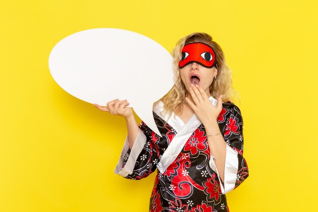 Vue de face jeune femme en robe de nuit tenant un énorme panneau blanc bâillant sur mur jaune sommeil nuit lit modèle fille couleur