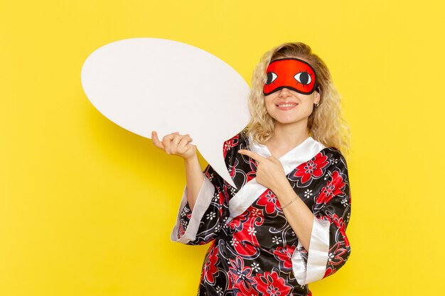 Vue de face jeune femme en robe de nuit tenant un énorme panneau blanc et souriant sur le mur jaune sommeil nuit modèle fille couleur