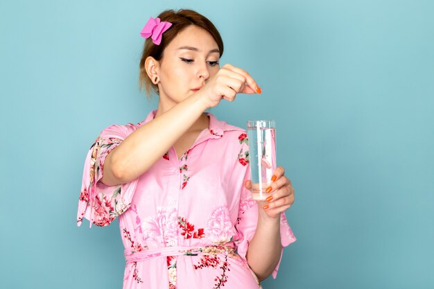 Une vue de face jeune femme en robe rose conçu de fleurs mettant la pilule dans le verre d'eau sur bleu