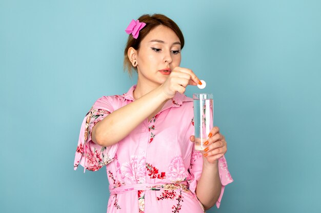 Une vue de face jeune femme en robe rose conçu de fleurs mettant la pilule dans le verre d'eau sur bleu