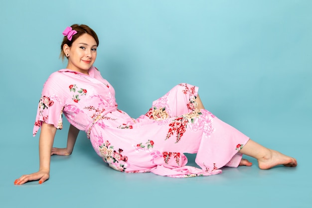 Une vue de face jeune femme en robe rose conçue de fleurs assis et posant avec sourire sur bleu