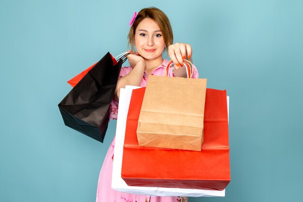 Une vue de face jeune femme en robe rose conçue de fleurs tenant des paquets d'achat et souriant sur bleu