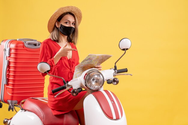 Vue de face jeune femme en robe rouge sur un cyclomoteur pointant sur la carte dans sa main