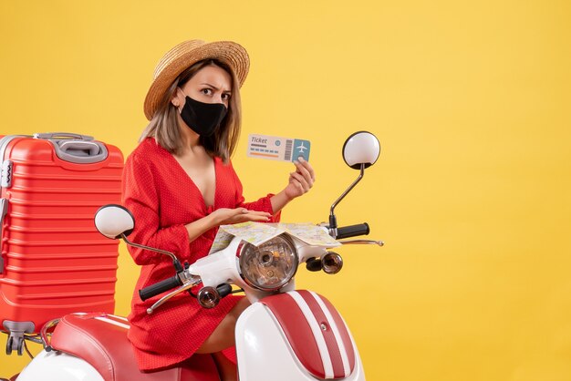 Vue de face jeune femme en robe rouge sur cyclomoteur avec valise tenant un billet