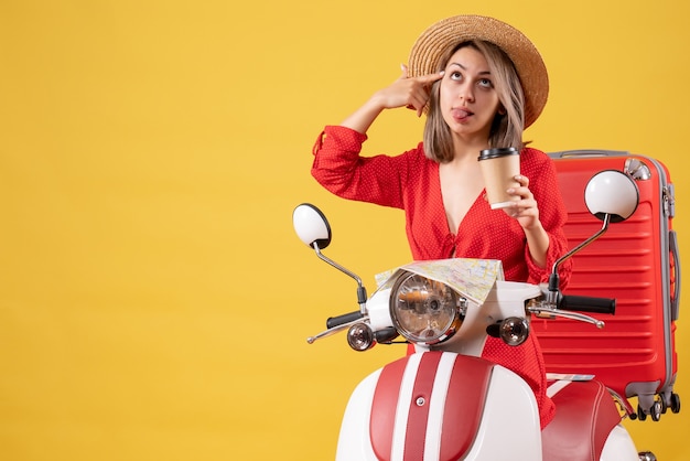 Vue de face jeune femme en robe rouge qui sort la langue tenant une tasse de café près d'un cyclomoteur