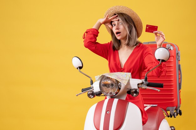 Vue de face jeune femme en robe rouge tenant une carte bancaire sur un cyclomoteur